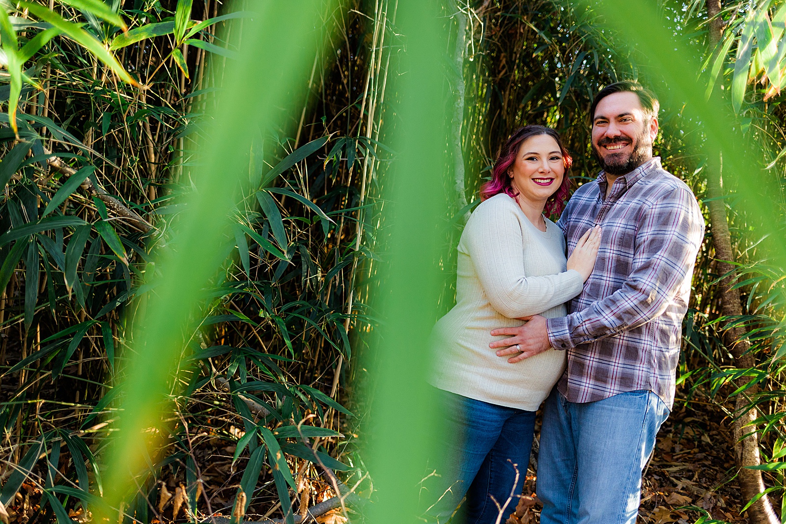 Couple smiles at the camera through some bamboo leaves in their neighborhood maternity session. 