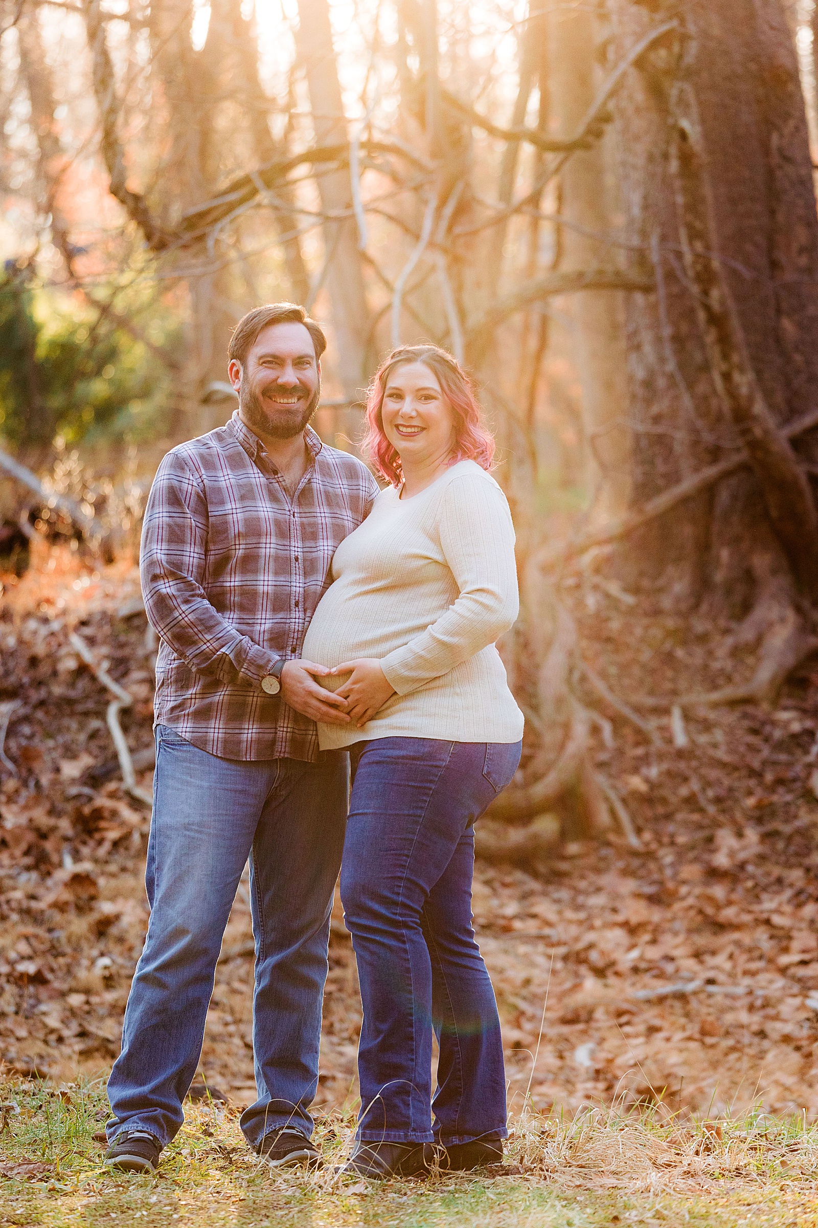 Sun shines through and couple smiles at the photographer while holding pregnant belly.