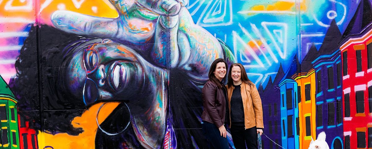 LGBTQ couple poses with their dog against a colorful mural for their Eckington DC engagement photography session.