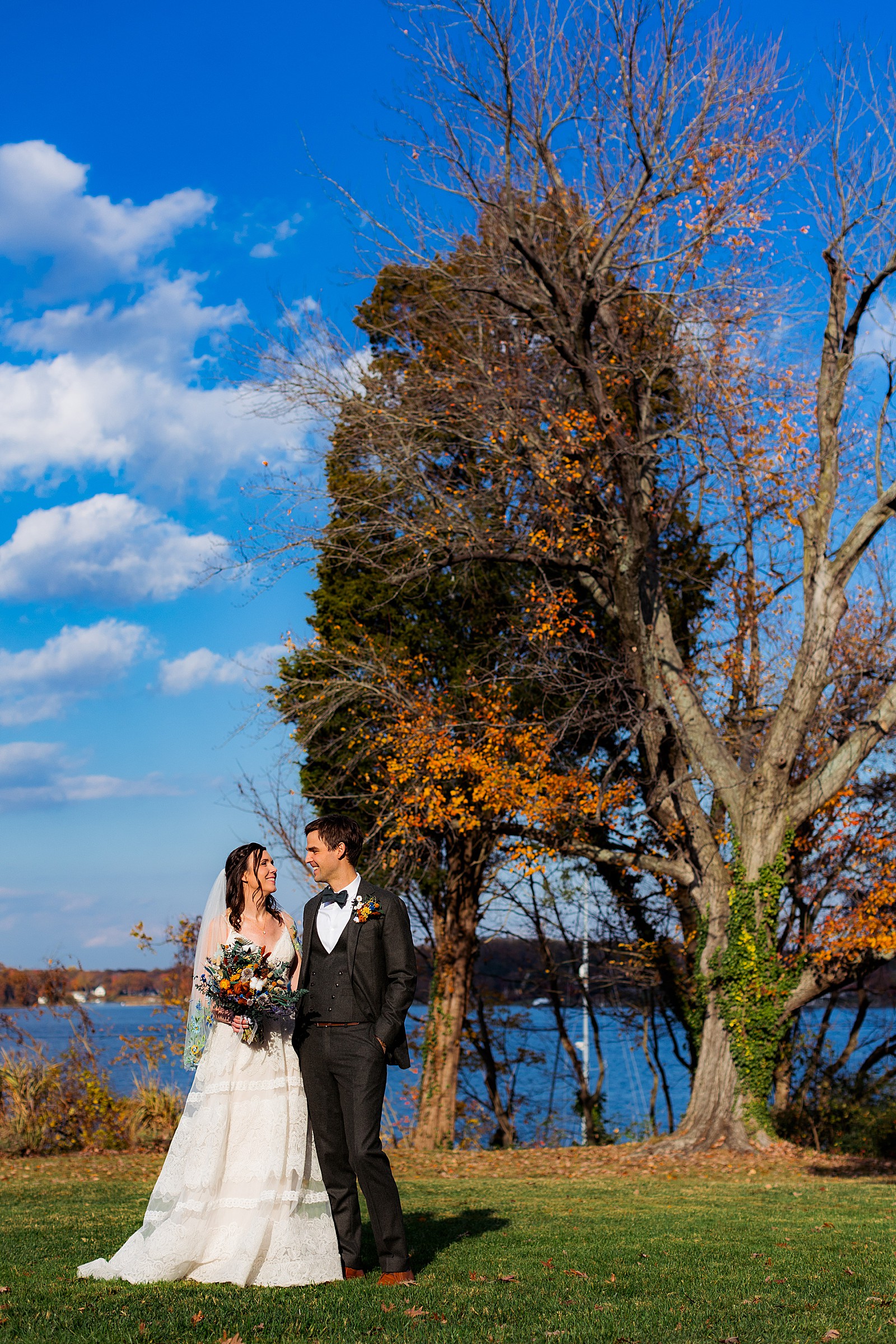 Bride and groom pose in front of some fall trees and the water at their fall London Town wedding.