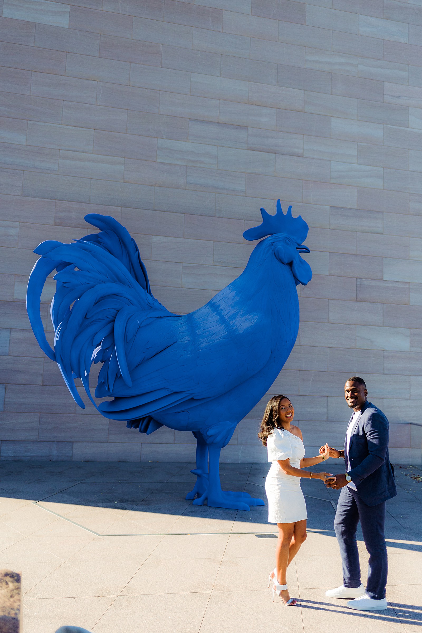 Playful couple dances in front of a blue rooster sculpture during their DC National Gallery of Art engagement photography session.