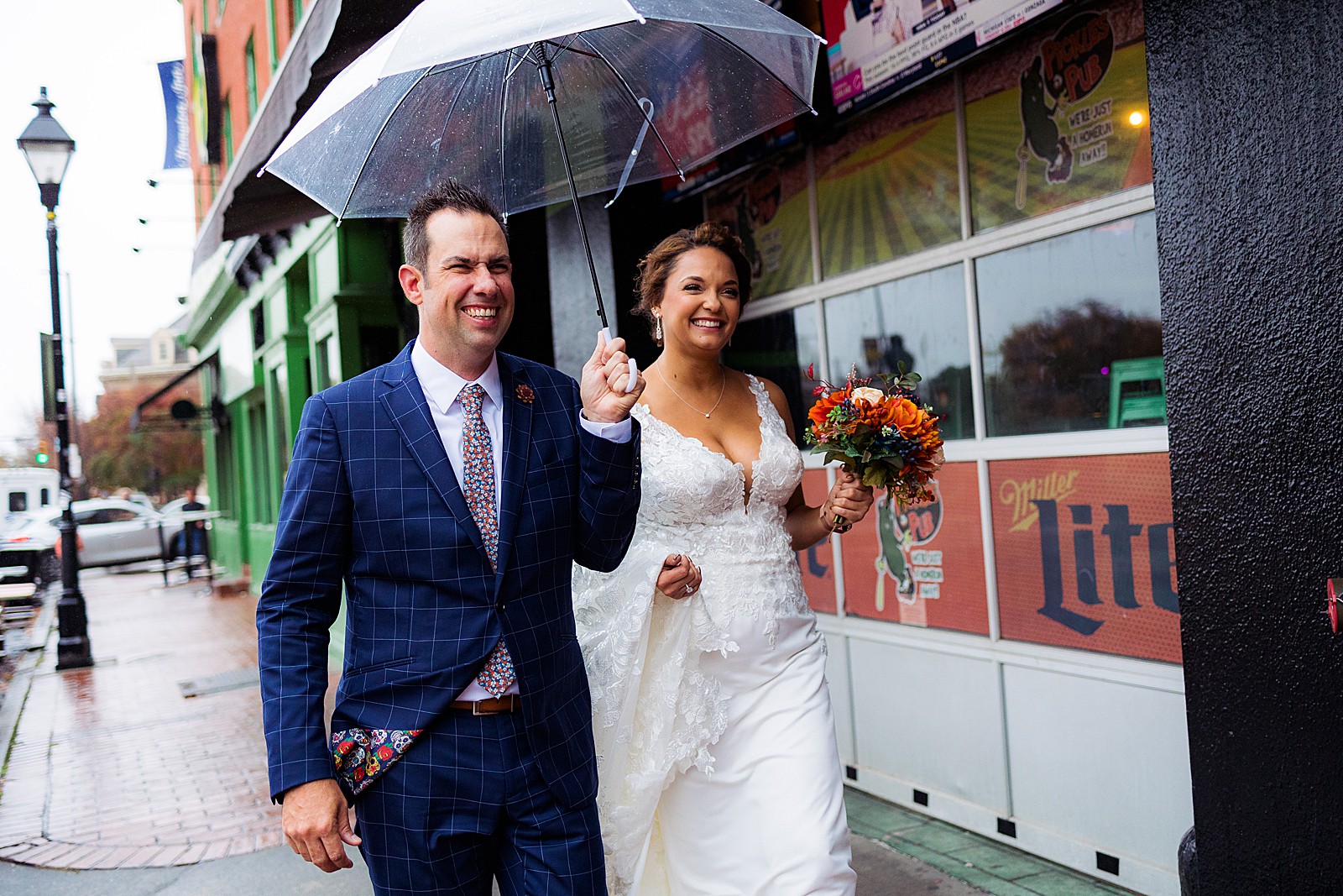 Bride and groom walk under an umbrella outside of Pickles Pub in Baltimore during their rainy day AVAM wedding.