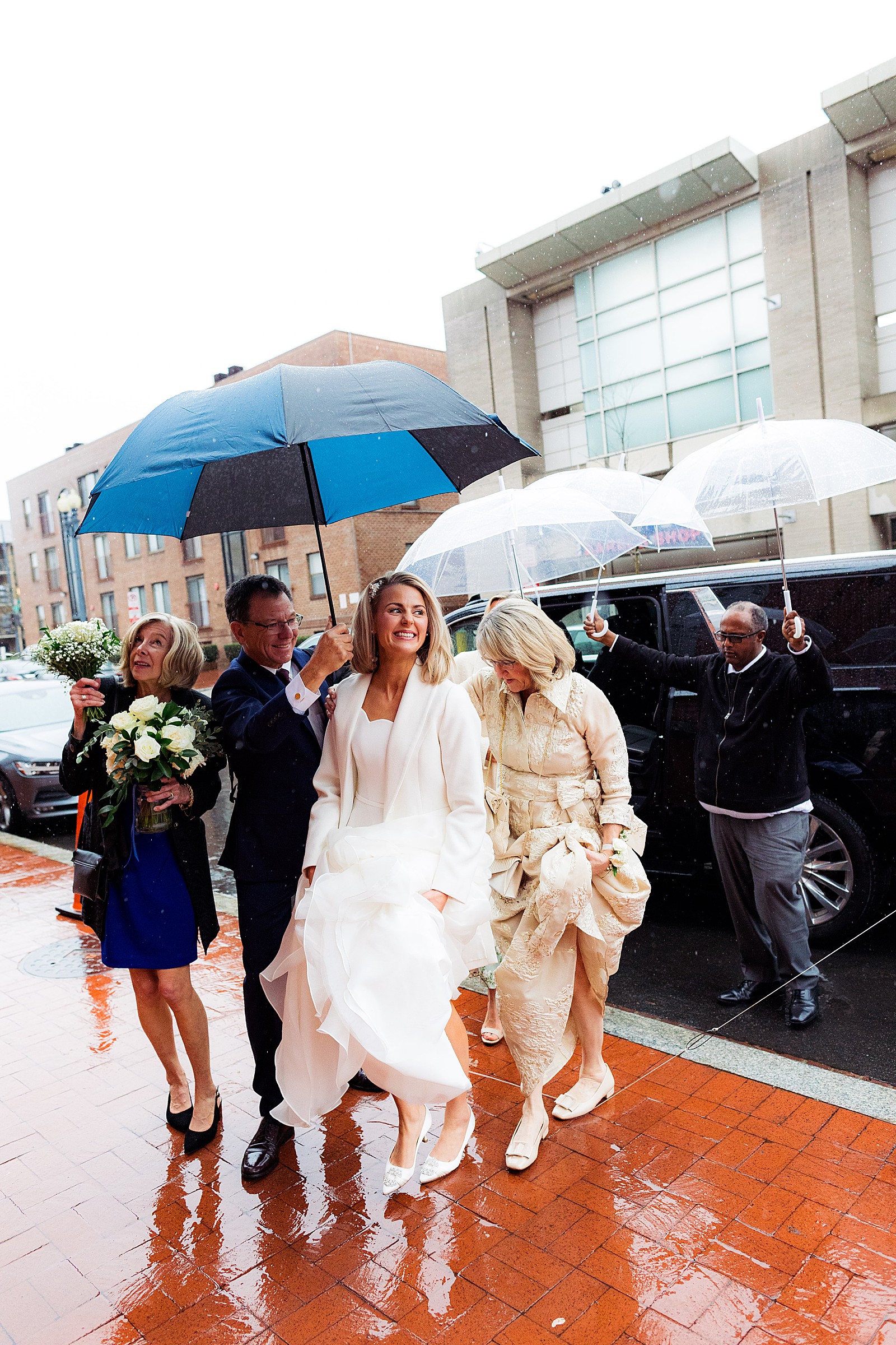 Bride gets escorted out of the car by multiple people holding umbrellas over her head on the way to her rainy wedding at Immaculate Conception church in DC.