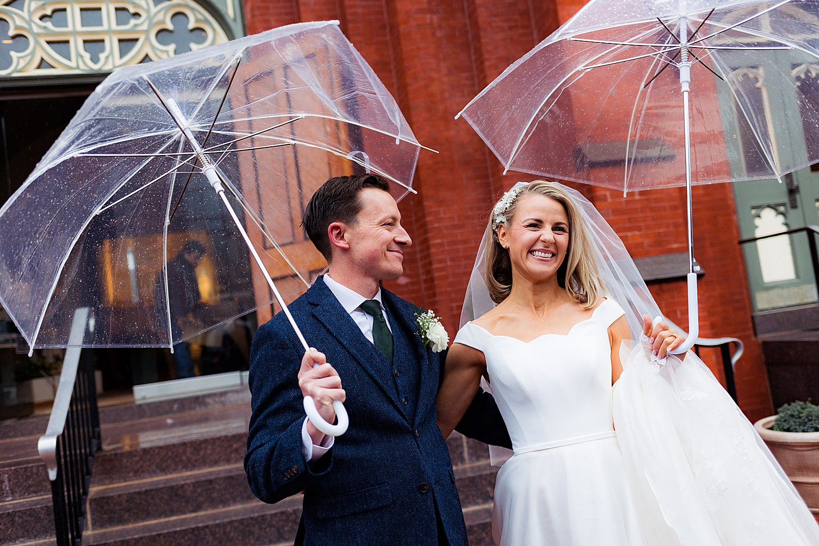 Bride and groom exit the Immaculate Conception church in DC and walk down the stairs with umbrellas in the rain.